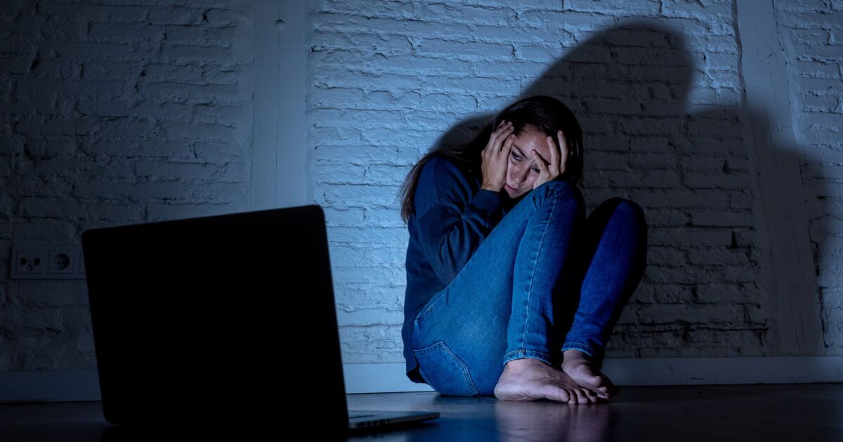 Young woman on the floor cowering against wall in dark room, lit only by the reflection from her laptop screen. She is looking fearfully at the laptop with her head in her hands.
