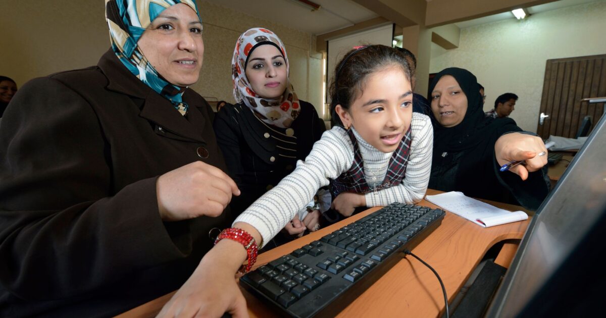 A girl reaches for a computer mouse in front two women in headscarves who are looking intently at a computer screen. A third woman in a headscarf points to something on the screen