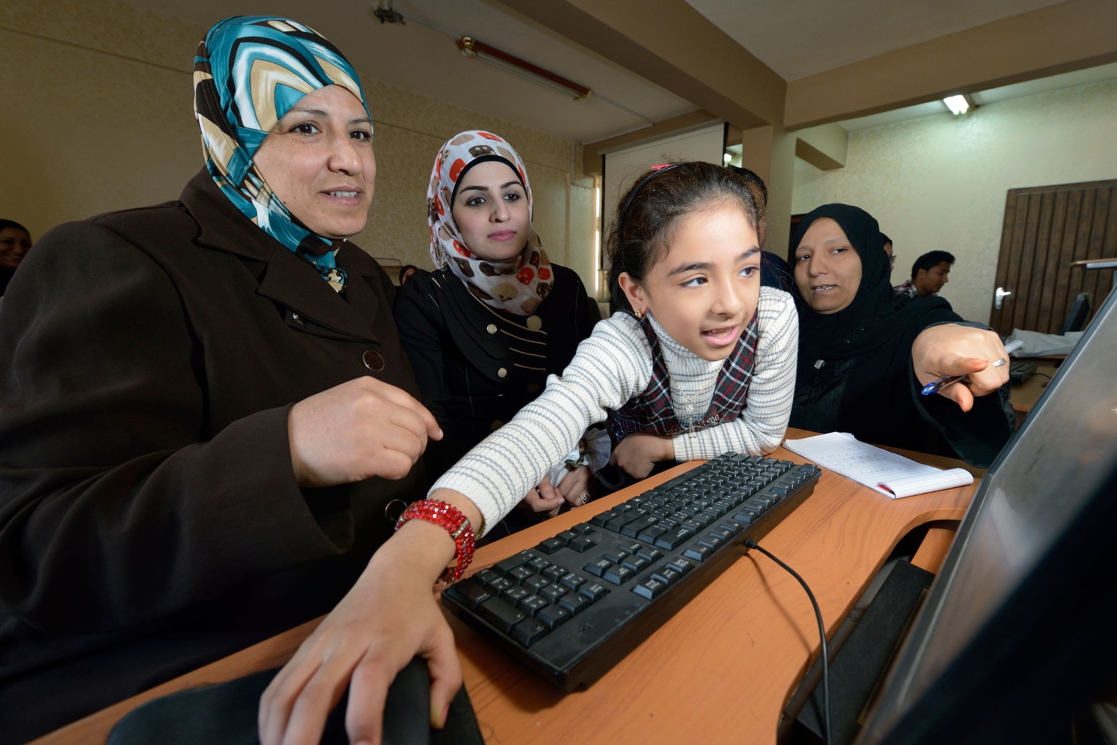 A girl reaches for a computer mouse in front two women in headscarves who are looking intently at a computer screen. A third woman in a headscarf points to something on the screen