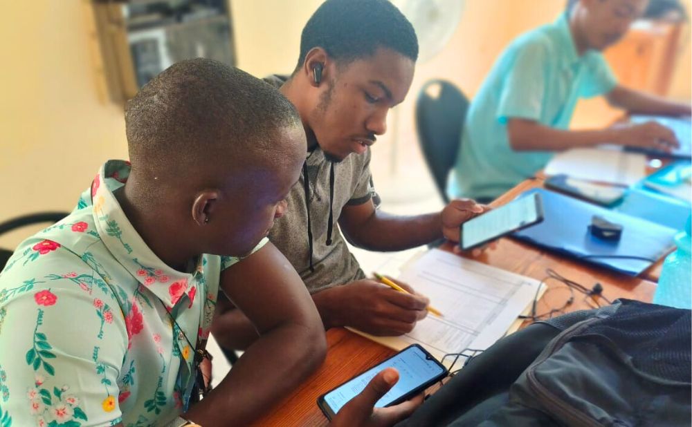 Two young Haitian men look at news on a mobile phone and make notes in a coding grid spreadsheet